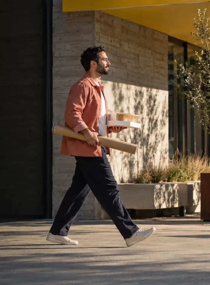 An architect in a coral jacket carries a plan tube and a small building model past his studio